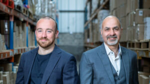 Two men stand in a warehouse filled with shelves of boxes, representing their green fulfillment initiative. Both are wearing suits and looking towards the camera. The background is blurred, focusing attention on the men.