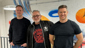 Three men, including Rob Morrice, stand in front of a vibrant mural on a brick wall, posing for a group photo. The man on the left and the man on the right wear black shirts, while Rob Morrice, in the middle, sports glasses and a graphic T-shirt.