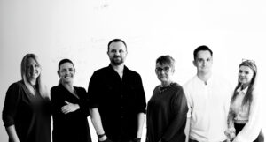 A group of six people standing in a row, posing for a photo against a white background at the PR Moment Awards. Four women and two men are looking at the camera, with some smiling and others maintaining neutral expressions.