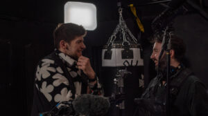 Two men stand smiling and talking amongst film equipment in a dimly lit setting. One wears a patterned jacket, and the other is equipped with audio gear. A light shines in the background, creating an ambiance perfect for their lively discussion on SEO strategies.