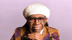 A person wearing glasses, a white hat, and a patterned jacket smiles with hands clasped together against a light purple background, embodying the spirit of the Leeds International Festival of Ideas.
