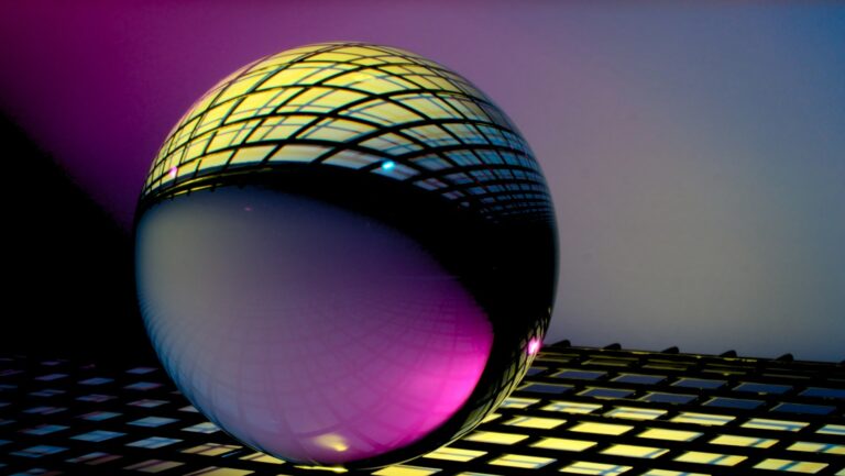 A reflective glass sphere sits on a grid-patterned surface, showcasing a colorful pattern of light in shades of purple, yellow, and blue. The scene feels almost otherworldly, as if crafted by AI against a gradient background.
