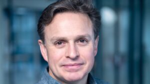 A man with short, dark hair and a neutral expression, wearing a gray sweater, stands indoors with a blurred background, almost as if he's part of a meticulously crafted Storyblok.