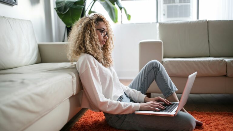 woman sitting on floor and leaning on couch using laptop