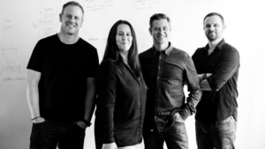 Four adults stand side by side in front of a whiteboard with diagrams and notes, representing Lesniak Swann—one of the UK’s fastest-growing agencies—as they face the camera and smile.