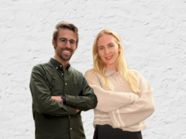 Two people standing side by side with arms crossed, smiling at the camera, in front of a white textured wall.