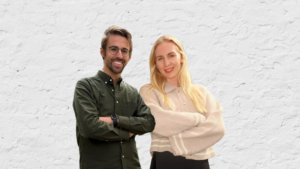 Two people standing side by side with arms crossed, smiling at the camera, in front of a white textured wall.