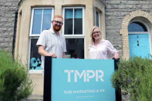 Two people, part of the senior team, stand outside a stone building, smiling and holding a sign that reads "TMPR B2B MARKETING & PR" with the website www.tmpr.co.uk.
