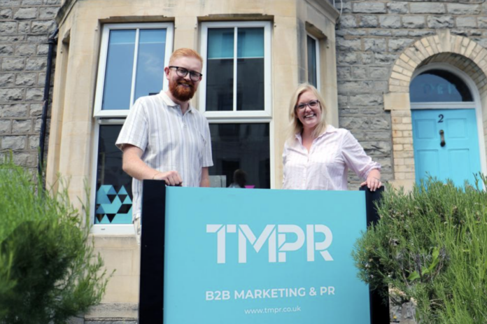 Two people, part of the senior team, stand outside a stone building, smiling and holding a sign that reads "TMPR B2B MARKETING & PR" with the website www.tmpr.co.uk.