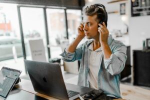 Multichannel sales: A person wearing headphones sits at a table with a laptop, smartphone, and papers, appearing focused in a well-lit modern workspace.