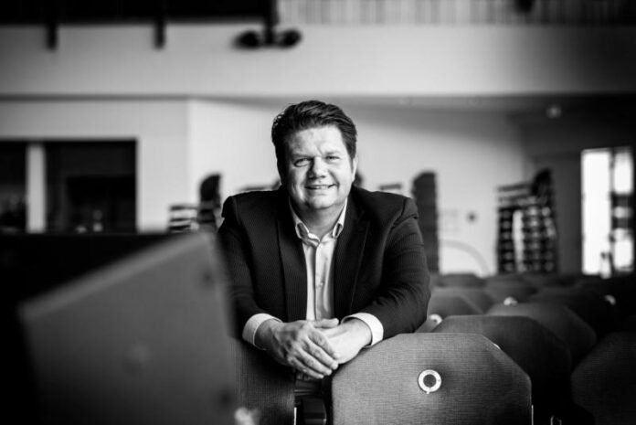 A man in a suit sits and smiles while leaning on the back of a chair in a large, empty indoor space. The photo is in black and white, reflecting Weitkamp marketing’s focus on German engineering SMEs seeking a visibility package.