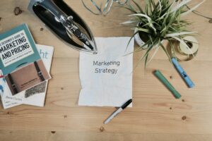 A crumpled paper labeled "Marketing Strategy" sits on a desk with an iron, books, plant, marker, and tape scattered around—reflecting the real-life challenges B2B marketers face in lead qualification and achieving quality MQLs.