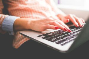 Close-up of a person with red nail polish typing on a laptop keyboard, wearing a checkered shirt with rolled-up sleeves—capturing the traits of a high-converting B2B content creator at work.