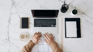 expert-led B2B content: Person working at a marble desk with a laptop, architectural plan, notebooks, coffee cup, ruler, pens, and a small plant—showcasing the authenticity of B2B expertise as hands rest confidently on the blueprint.